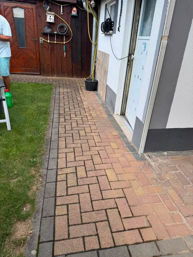 Pflasterweg an der Hauswand vor der Reinigung, mit dunklem Belag und Bewuchs in den Fugen.